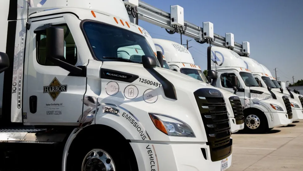 Electric semi trucks parked at a charging station with overhead charging equipment, representing challenges in heavy-duty EV infrastructure deployment.