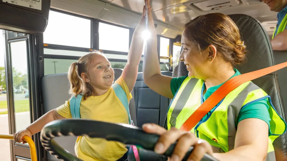 A school bus driver wearing a safety vest exchanges a high-five with a smiling young student near the front of a school bus. Sunlight streams through the windows as another adult stands nearby.