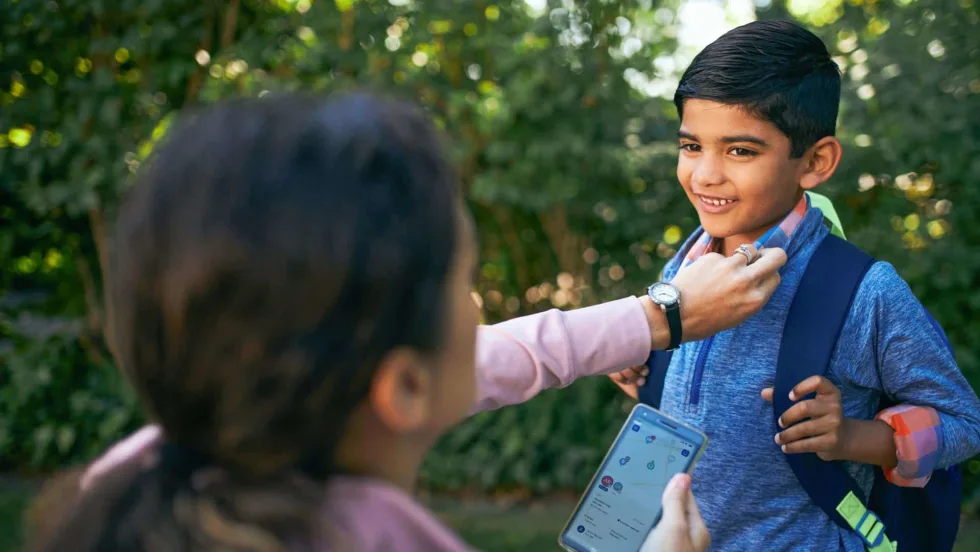 A parent uses the FirstView mobile app while helping a student with his backpack, illustrating real-time school transportation tracking and communication.