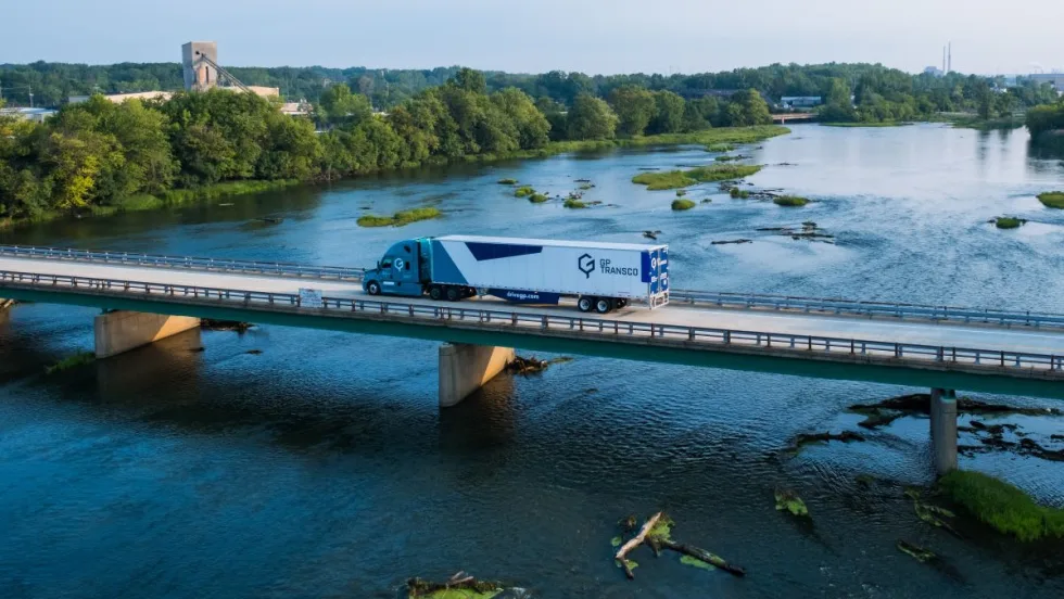 A GP Transco tractor-trailer crosses a bridge over a river, with trailer side skirts visible beneath the white trailer.