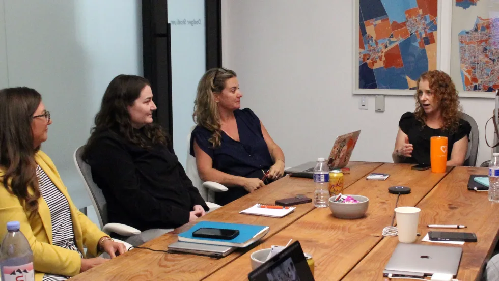 Four women participate in a council meeting around a wooden conference table, with one speaking while the others listen. Laptops, notebooks, and drinks sit on the table.
