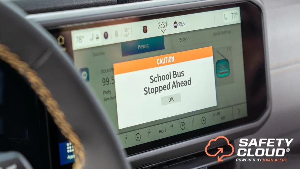 Vehicle dashboard display showing a “Caution: School Bus Stopped Ahead” warning from HAAS Alert’s Safety Cloud system, designed to alert motorists to stopped school buses.