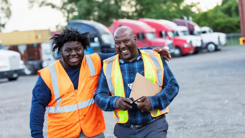 Two black men in safety vests walking together laughing in a truck fleet yard