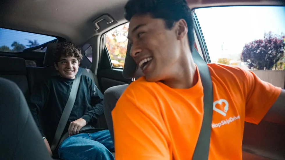 HopSkipDrive driver in an orange shirt smiles at a seat-belted student riding in the back seat of a car.