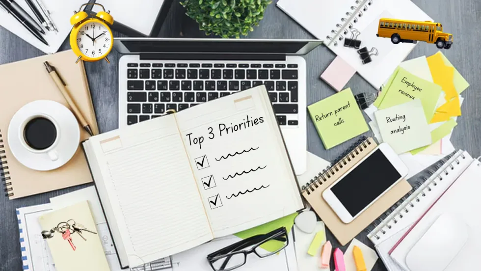 Overhead view of a busy workspace with a laptop, notebooks, coffee, phone, and scattered sticky notes labeled tasks like “Return parent calls” and “Routing analysis,” centered around an open notebook listing “Top 3 Priorities,” symbolizing time management and organization for a transportation supervisor