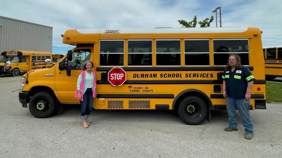 A Durham School Services bus with two people posing in front of it.