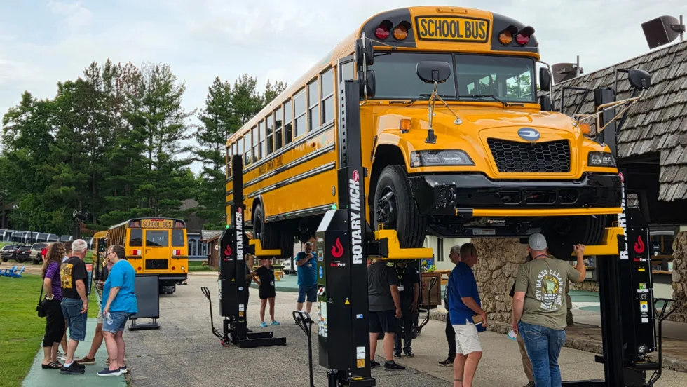 A new Blue Bird school bus is lifted on a heavy-duty Rotary vehicle hoist during an outdoor maintenance equipment demonstration as attendees gather nearby.