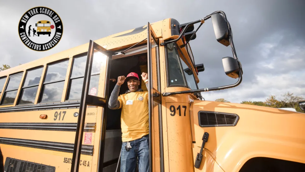 School bus driver stands in the doorway of a yellow school bus during the New York School Bus Contractors Association roadeo event, with dark storm clouds in the background and the NYSBCA logo in the corner.
