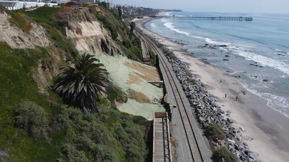 Coastal railroad tracks run between a steep bluff and the Pacific Ocean in San Clemente, California, with rock barriers protecting portions of the shoreline.