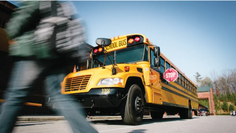 A yellow school bus with its red stop arm extended is stopped to load or unload students as a blurred child crosses in the foreground, illustrating the importance of motorists stopping for school buses and protecting students in the loading zone.