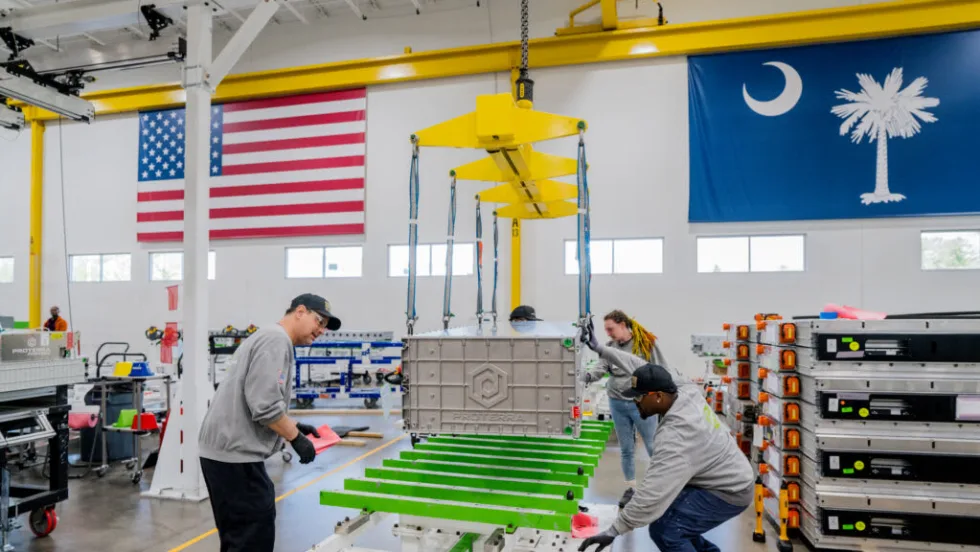 Workers assemble a large Proterra EV battery pack inside a manufacturing facility, using an overhead crane to position the battery module onto a chassis frame. American and South Carolina state flags hang above the production floor, with additional battery packs stacked nearby.