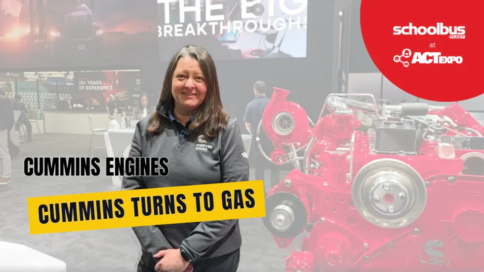 Andrea Lukas of Cummins stands beside a large red engine display at ACT Expo, with on-screen text reading “Cummins Turns to Gas” during a School Bus Fleet interview about new gasoline engine technology.