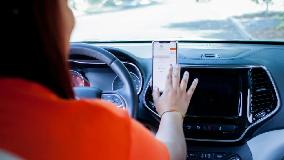 A driver in an orange t-shirt uses a smartphone mounted on a vehicle dashboard to access HopSkipDrive’s app.