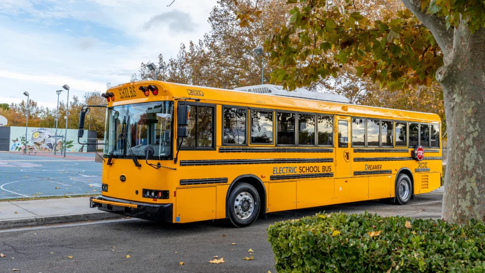 A yellow RIDE Type D electric school bus is parked along a tree-lined street outside a school.