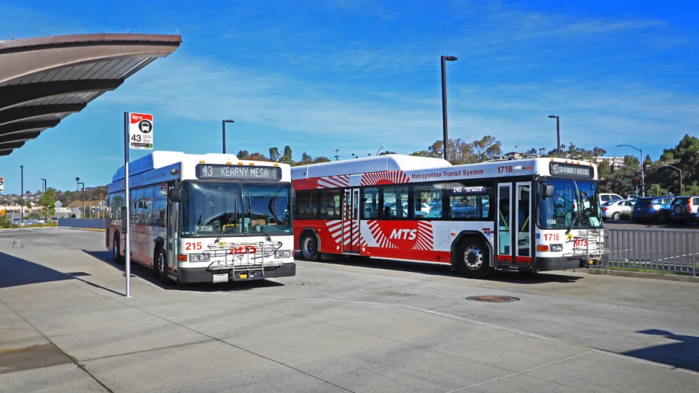 Two San Diego Metropolitan Transit System buses sit at a transit station under a clear blue sky.