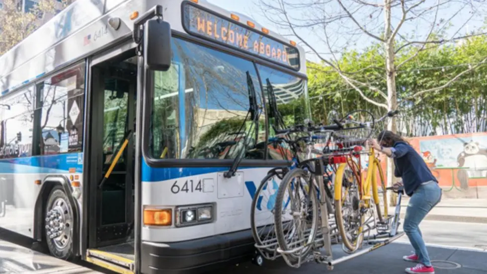 A passenger loads a bicycle onto the front bike rack of a VTA transit bus stopped along a city street.