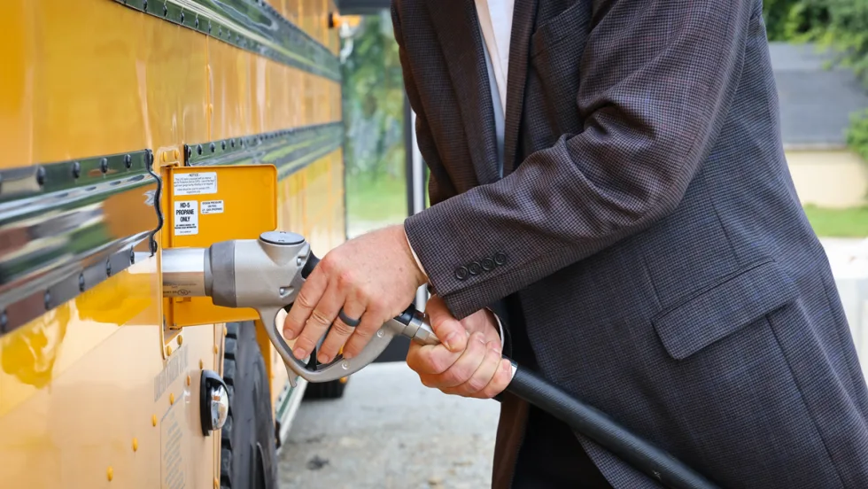 Close-up of a person refueling a yellow school bus with propane. The fuel nozzle is connected to the bus's propane fueling port, illustrating an alternative fuel option for school transportation fleets.