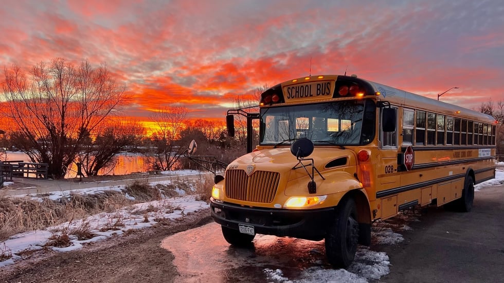 A yellow school bus is parked beside a snow-lined roadway at sunset, with vivid orange and pink clouds reflecting on a nearby pond. The bus's headlights and warning lights are illuminated against the colorful winter sky.
