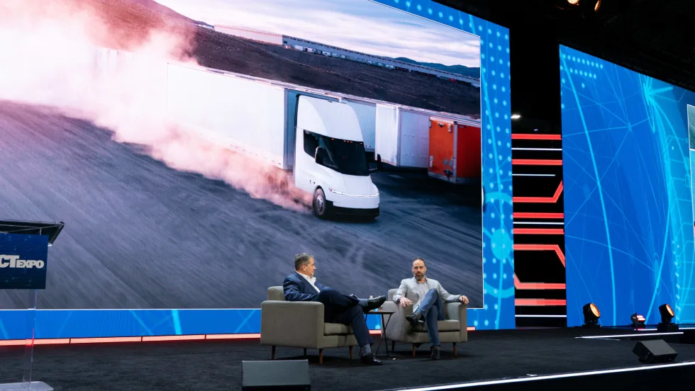 Two men in chairs on stage with big video screen behind them showing Tesla Semi