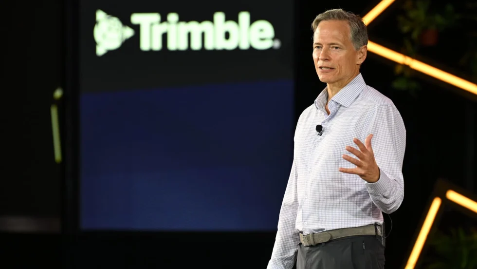 Photo of man in collared buttoned shirt speaking on stage in front of a backdrop that says Trimble