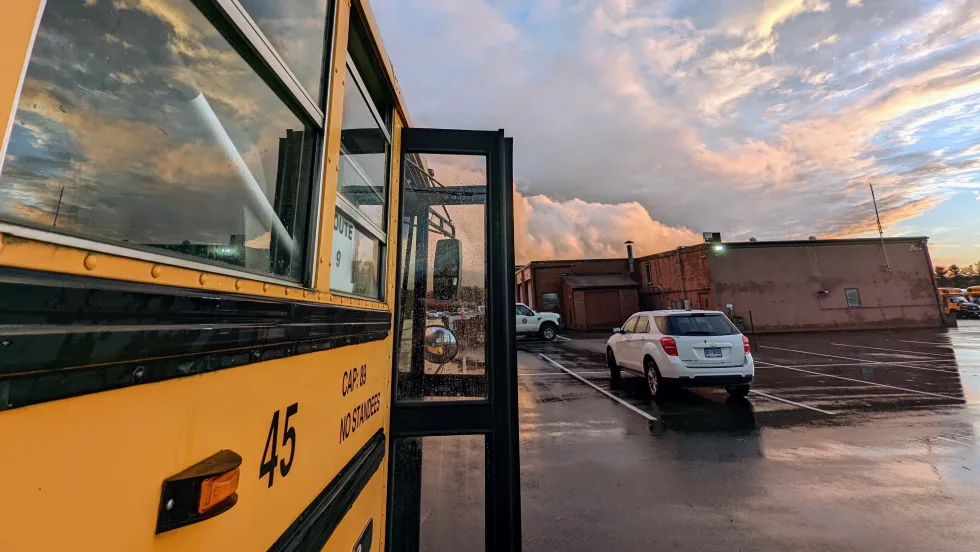 A yellow school bus with its service door open is parked after a rainstorm as dramatic clouds fill the evening sky. Reflections shimmer in the wet pavement, creating a scenic view of the bus yard at sunset.