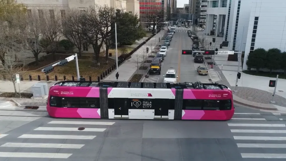 An pink and white OKC streetcar moves down a street. 