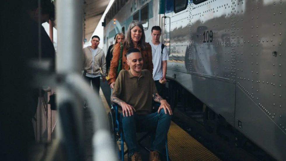 A woman pushes a man in a wheelchair on a rail platform.