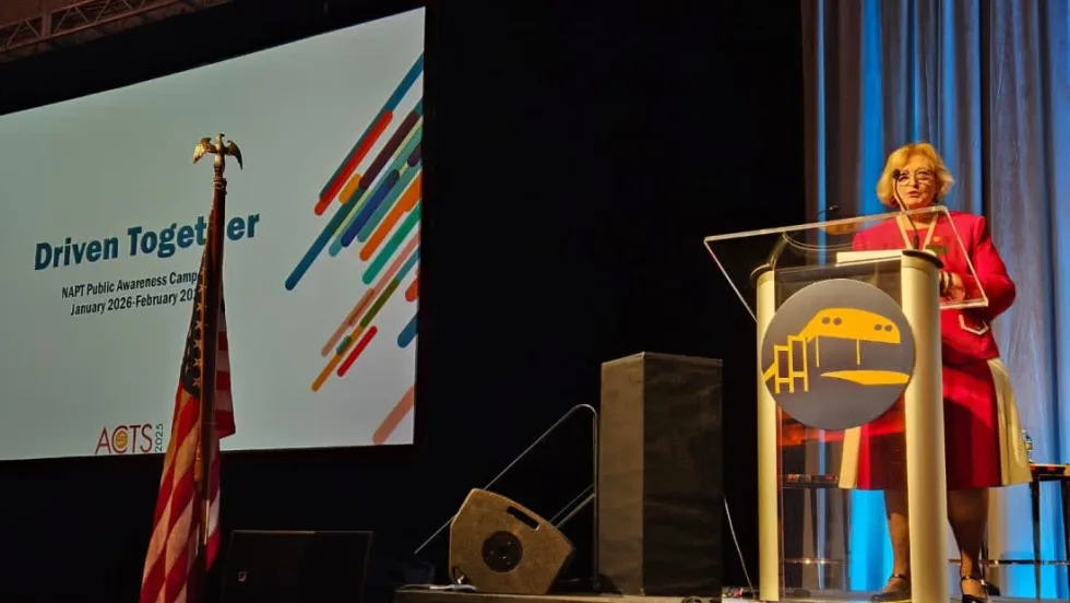 Molly McGee Hewitt stands at a podium on a stage with a screen behind her that says "Driven Together" and the NAPT logo