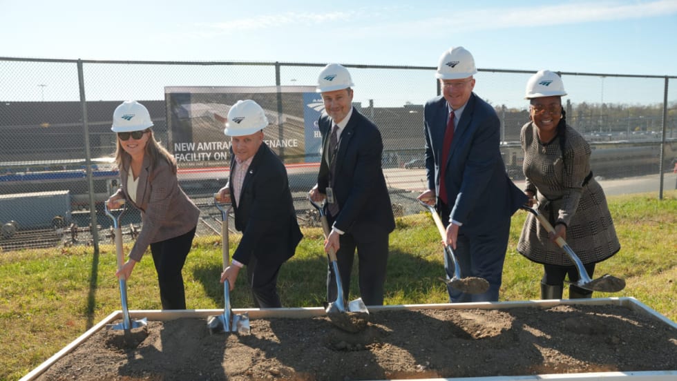 Five people in business attire lined up shoveling dirt.