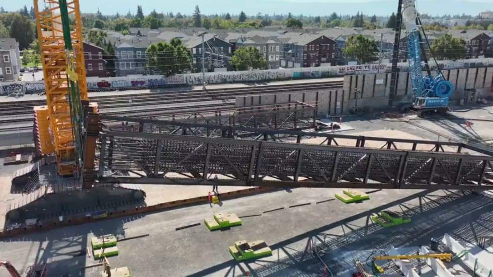 A large yellow crane lifts a steel rebar cage on one side.
