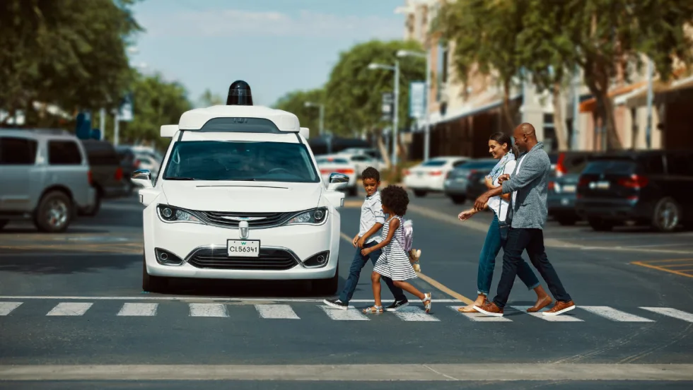 A white Waymo vehicle waits at a crosswalk as a family crosses.