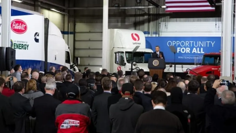 President Obama delivers remarks before a crowd in a large distribution center with Safeway trucks and an American flag in the background.