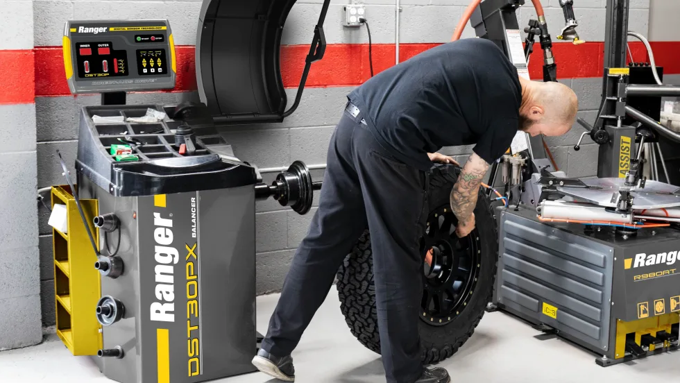 Automotive technician handling a large tire and wheel assembly beside a Ranger DST30PX wheel balancer and tire-changing equipment in a professional repair shop.