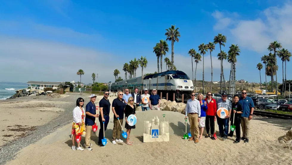 A group of people stand on a beach with a rail car in the background.