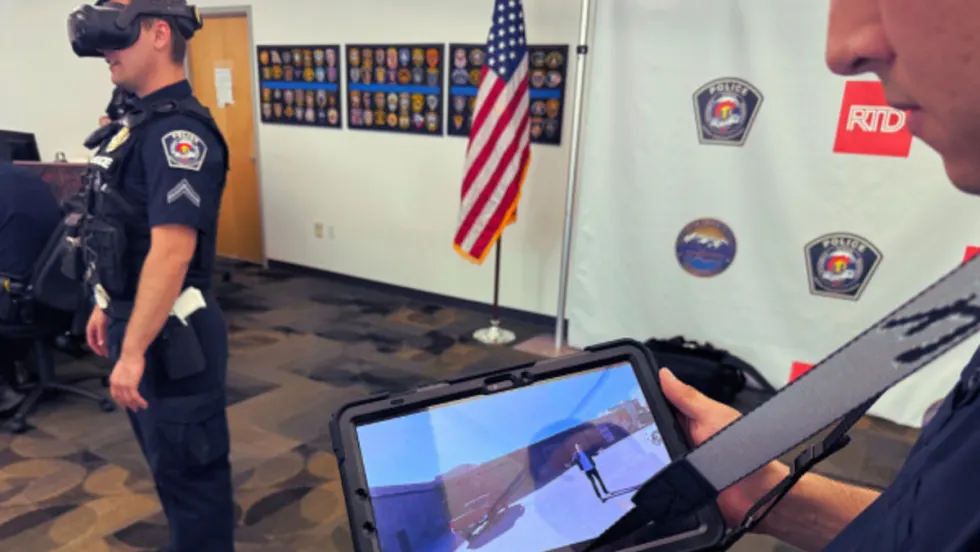 A RTD transit police offer holds up a tablet showing virtual reality training while an officer in the background stands with a VR headset on.