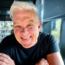Smiling man with white hair wearing a black shirt and sitting with his arms crossed at a table.