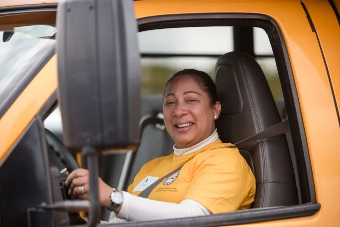 Woman wearing a New York State School Bus Safety Competition shirt smiles from the driver’s seat of a yellow school bus while buckled in behind the wheel.