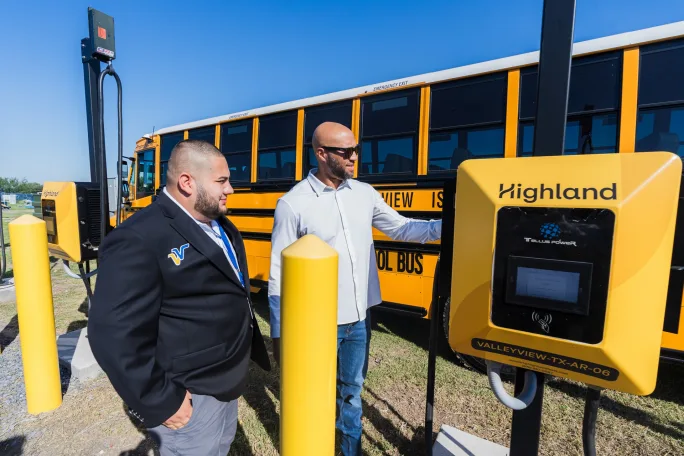 Two people stand near an electric school bus charger at Valley View ISD, discussing the new Highland Electric Fleets charging system installed for the district’s electric buses.