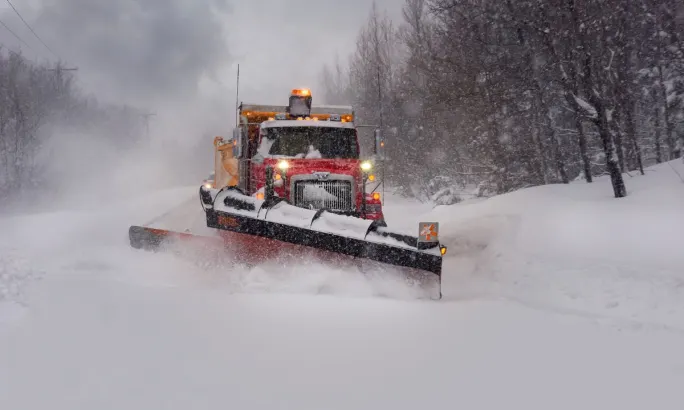 A red snowplow truck clears a snow-covered road during heavy snowfall, with headlights and warning lights on, pushing snow to the side while driving through a wooded area.
