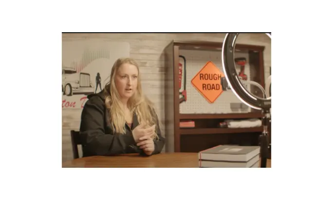 A woman with long blonde hair speaks while seated at a table, with a “Rough Road” sign and truck-themed decor behind her, featured in the ATA’s DRIVEN series on professional truck drivers.