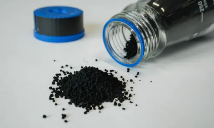 A small pile of black granular material spilled from a clear lab jar with a blue rim, lying on its side on a white surface. The blue cap is placed nearby in the background.