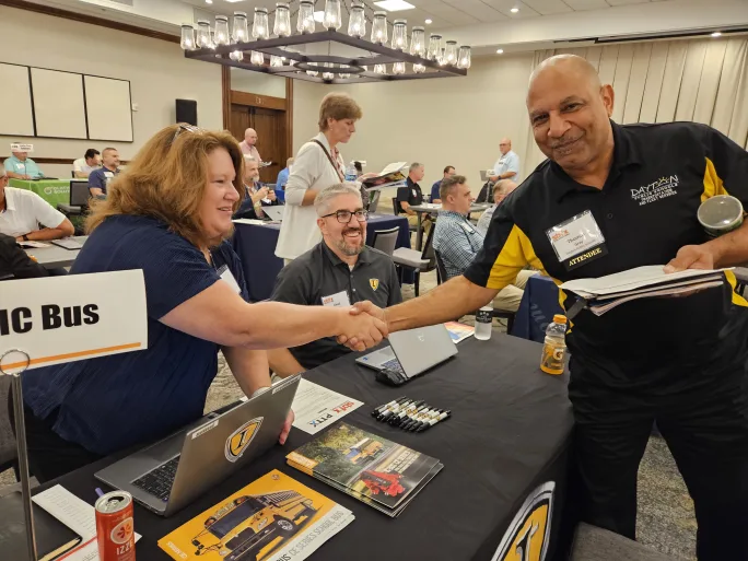 A man and a woman shakes hands across a table in a large room during an event.