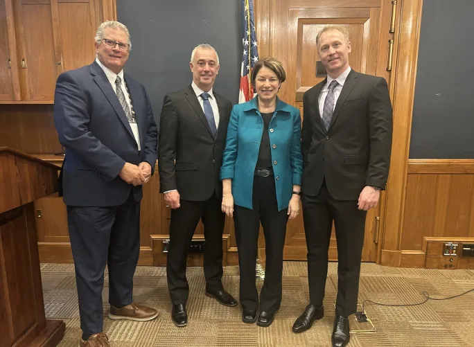 Three men in black suits stand with Amy Klobuchar in the capitol building