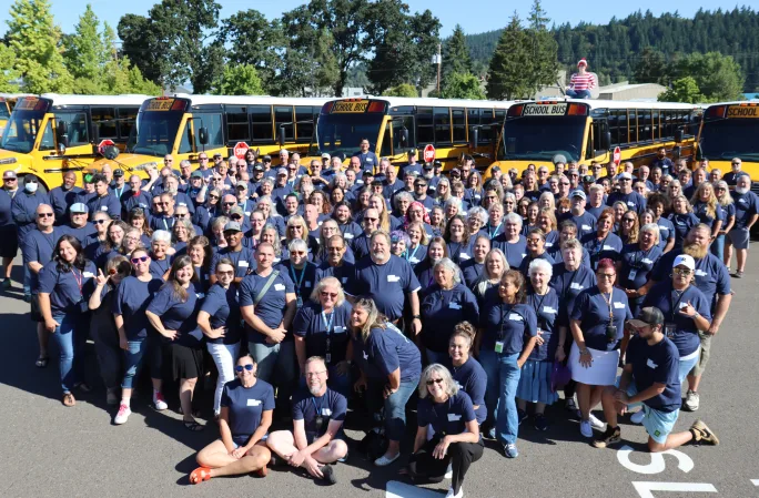 a large group of people pose together in front of a row of school buses all wearing an NCSD navy blue t-shirt