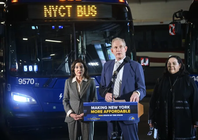 Three people stand behind a podium placed in front of a NYCT bus.