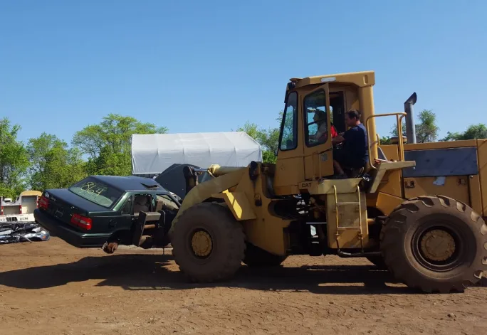 Wheel loader moves a stripped vehicle through an automotive recycling yard. Crushed vehicles, scrap metal, and dismantled parts are visible in the background under a clear blue sky.