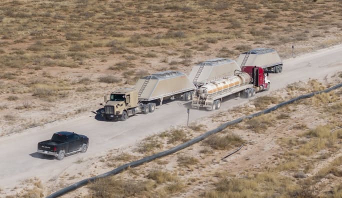 A Kodiak-powered autonomous truck hauling triple trailers of frac sand travels on an oilfield road in the Permian Basin alongside other vehicles.
