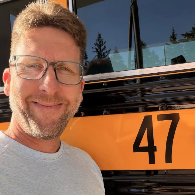A man wearing glasses and a gray T-shirt smiles for a selfie beside a yellow school bus marked with the number 47.