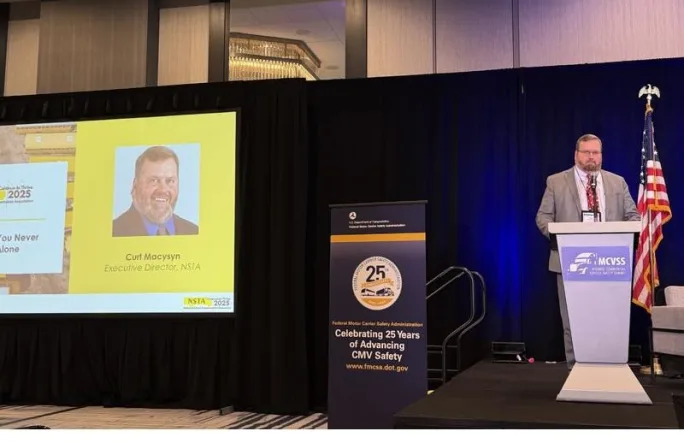 Curt Macysyn stands on stage at the MCVSS event giving a presentation. Behind him is a presentation screen with his photo, title, and company listed, and the podium has a blue event logo.
