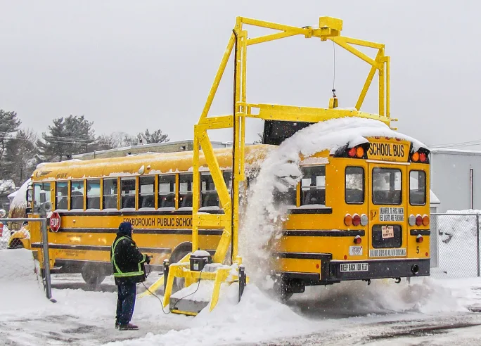 a school bus drivers under a snow scraper mounted to the ground outside a bus shop